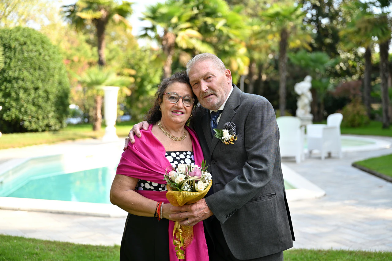 Salvatore e Maria sorridono abbracciati nel giardino accanto alla piscina, durante il loro anniversario di matrimonio.